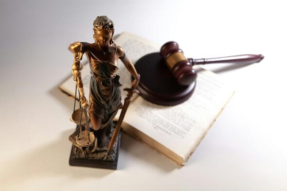 Bronze Lady Justice statue holding scales stands beside wooden gavel and open law book on desk symbolizing fairness and rights for those who are legally blind. Close perspective highlights balance and accessibility in legal system.