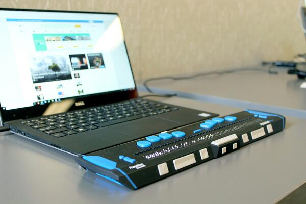 Laptop connected to a brallie display device with tactile buttons and refreshable braille cells for visually impaired users working on digital content.