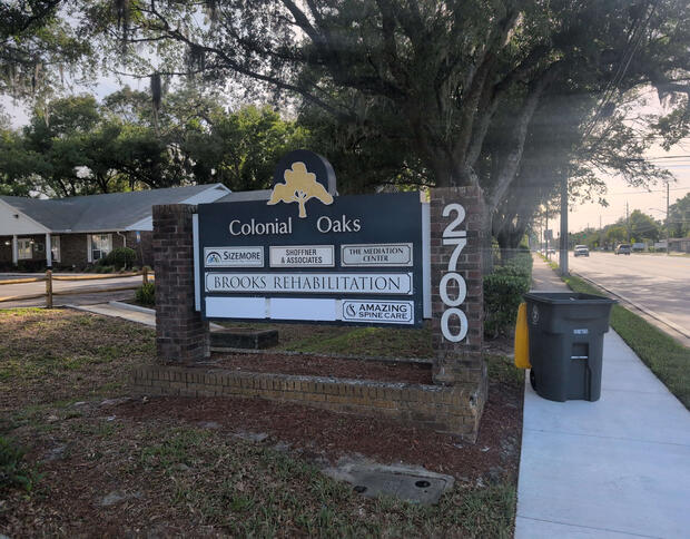 Alt text: Roadside sign in Jacksonville FL reads "Colonial Oaks", "Brooks Rehabilitation", "Sizemore", "Shoffner & Associates", "The Mediation Center", and "Amazing Spine Care" near sidewalk.