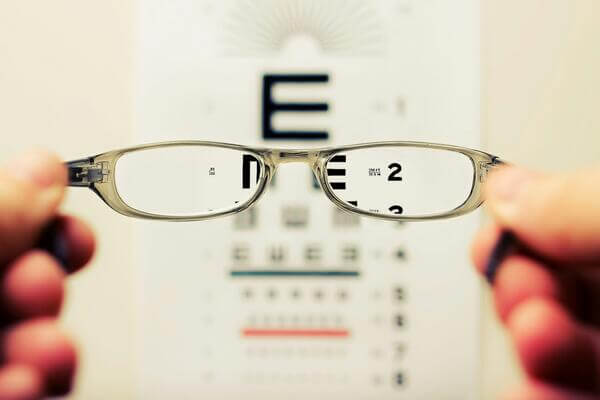 Hands hold eyeglasses in front of a blurred eye chart, where letters become clear through the lenses, demonstrating vision correction for people with visual impairment.